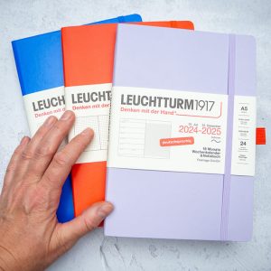 Jemand legt verschiedenfarbige Kalender, Planer und Notizbücher von LEUCHTTURM1917 auf einem grauen Schreibtisch aus.