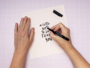 Jemand lettert mit einem schwarzen Kreativmarker von Faber-Castell den Satz „a little note to say thank you“ auf ein quadratisches Stück rosa Fotokarton. 