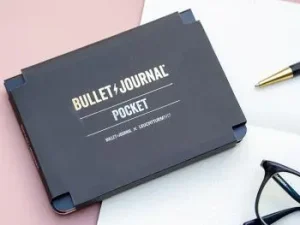 Das Produkt Bullet Journal Pocket von LEUCHTTURM1917 liegt auf einer rosa Schreibtischunterlage. Daneben sind eine Brille und ein Drehgriffel zu sehen. Foto: