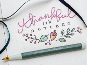 Monatsdeckblatt für Oktober in einem Bullet Journal von LEUCHTTURM1917. Das Motiv: Der Schriftzug „thankful it´s october“, darunter ein Doodle mit Herbstmotiven.