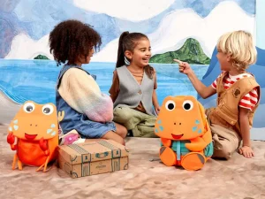 Drei Kindergartenkinder sitzen mit ihren Rucksäcken in Tierform von Affenzahn im Sand und reden miteinander.