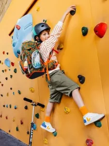 Ein Junge im Grundschulalter klettert mit seinem Schulranzen von ergobag eine orangefarbenen Boulderwand hoch.