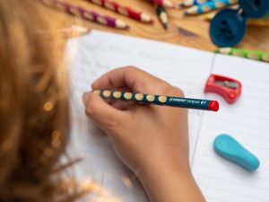 Ein Kind schreibt mit einem EASYgraph Bleistift von STABILO ein Diktat in ein Heft. Drumherum liegen Buntstifte, Spitzer und ein Radiergummi. Foto: Johanna Rundel