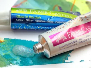 Eine Glitzer- und eine Glow-in-the-Dark-Paste von Faber-Castell liegen auf einem Tape-Resist-Art-Kunstwerk. Foto: Johanna Rundel