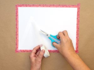 Jemand klebt wiederablösbares Klebeband auf ein weißes Blatt Papier auf einem mit Packpapier belegten Tisch. Foto: Johanna Rundel